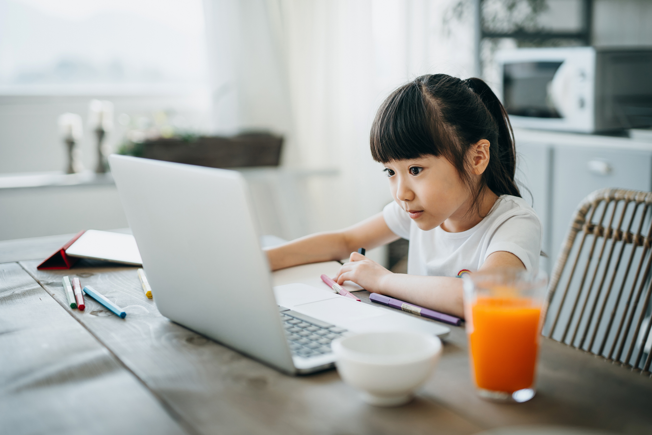 Little Asian girl studying from home. She is attending online school classes with laptop and digital tablet and writing notes at home