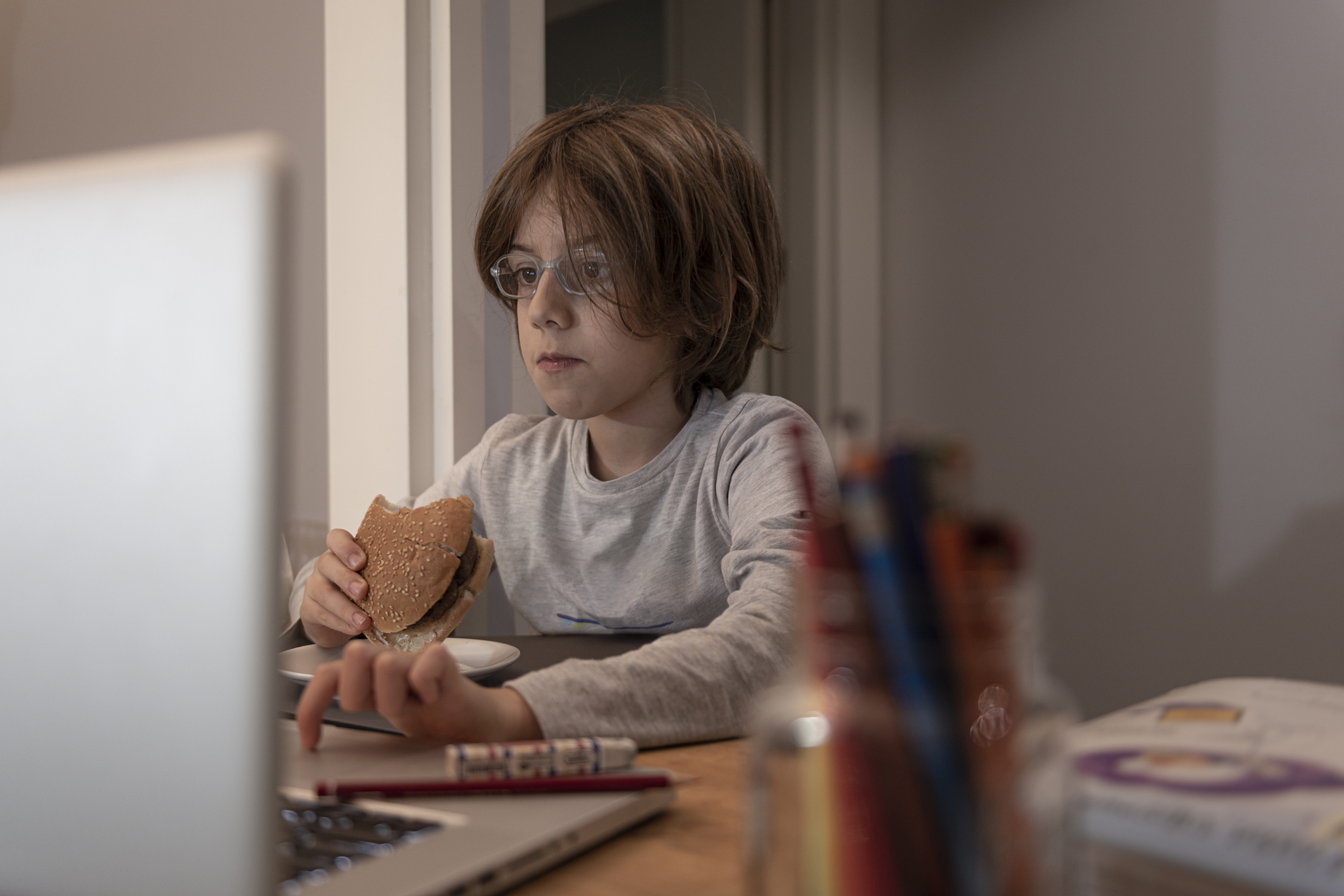 Little boy eating hamburger and during COVID-19 quarantine attending to online school class from his room.