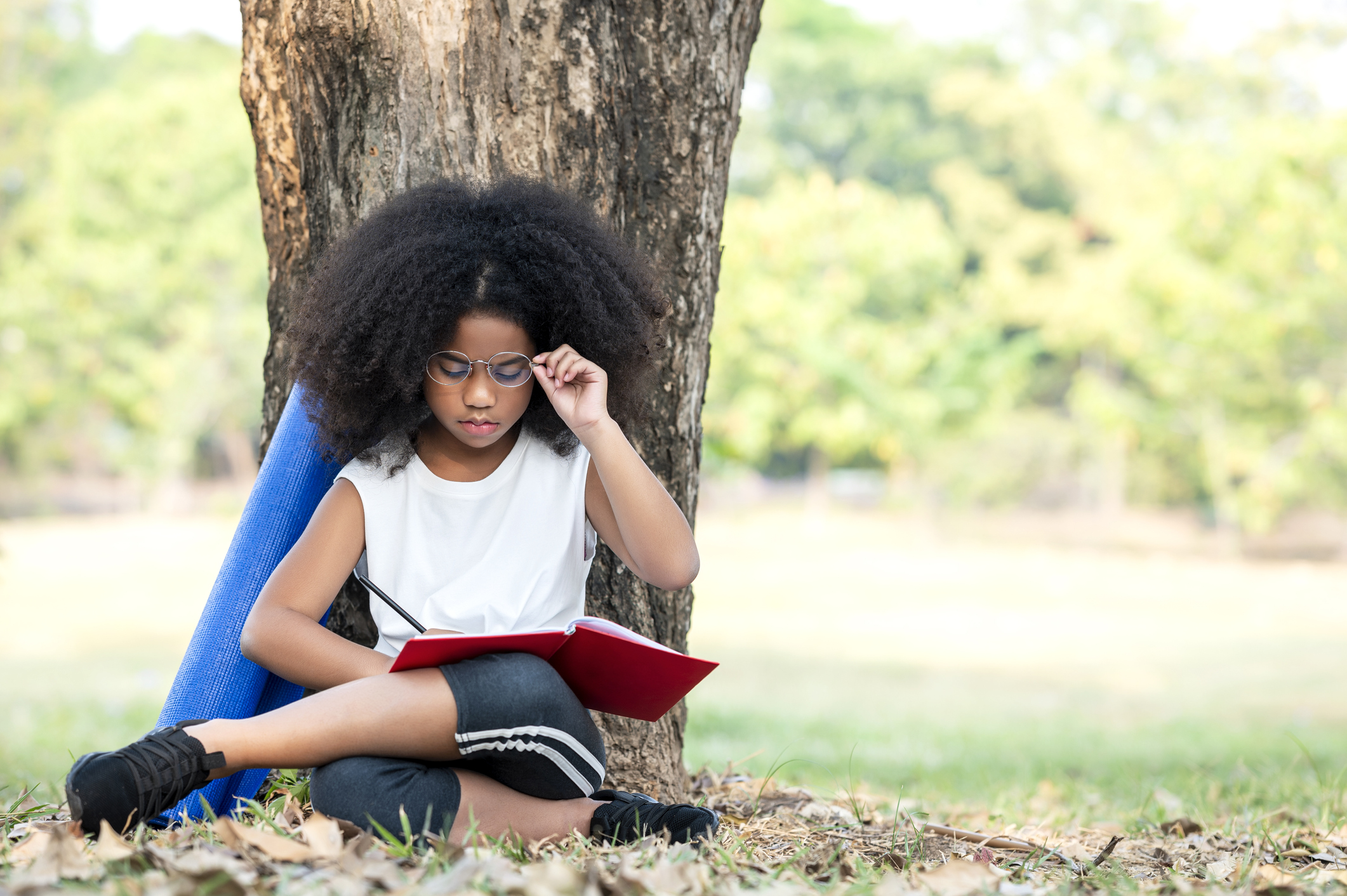 Afro curly hair teenager reading book at park while sitting leaning at a trees and meadow in the park. Feeling relax and comfortable, Learning, Leisure activity in park.