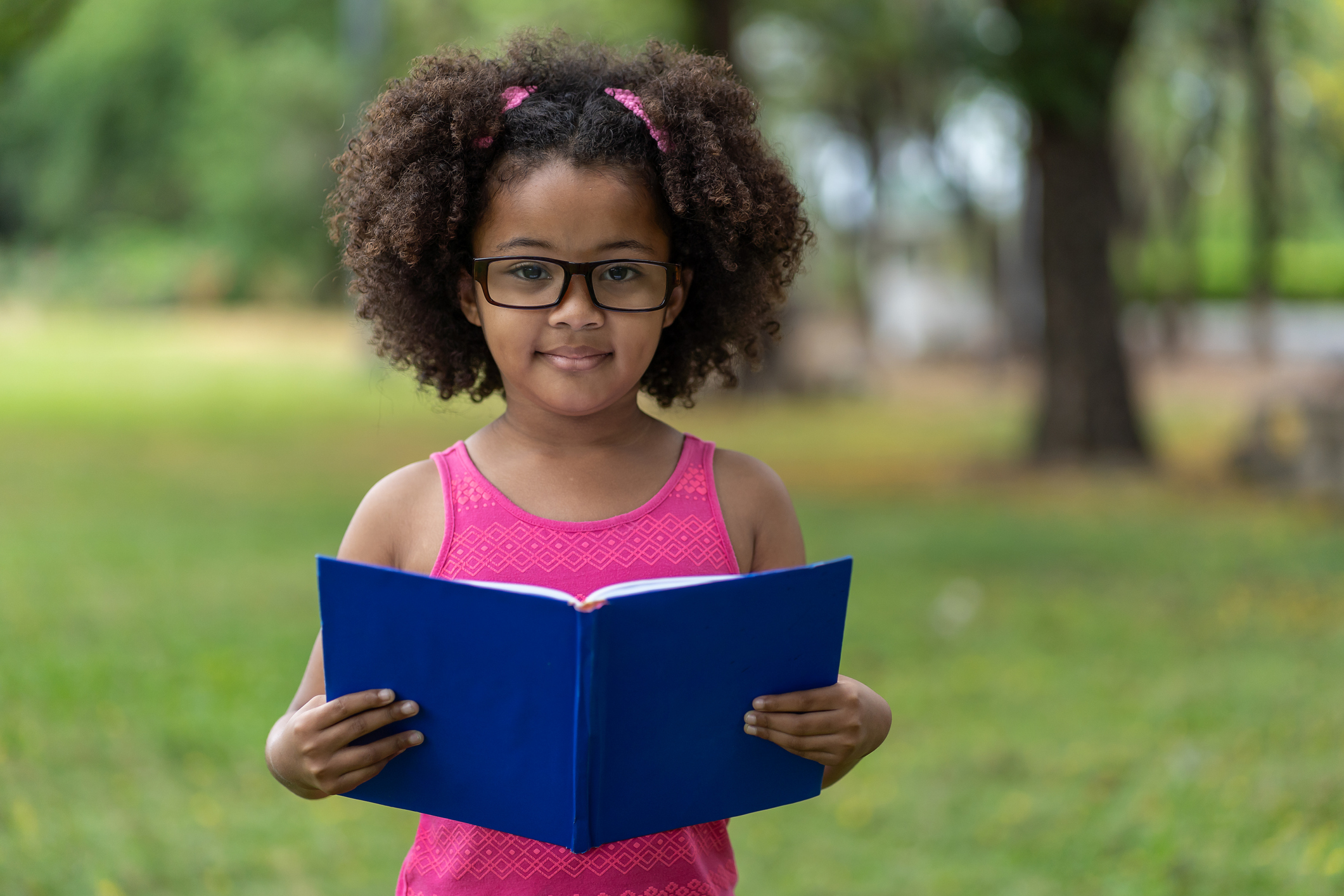 African American little girl withÂ curly and hair wearing glasses while reading a book at summer outdoor. Education or Field trip concept