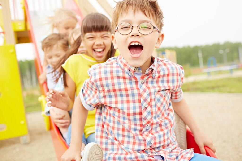 Boy with glasses leads group of children on a slide in a playground.
