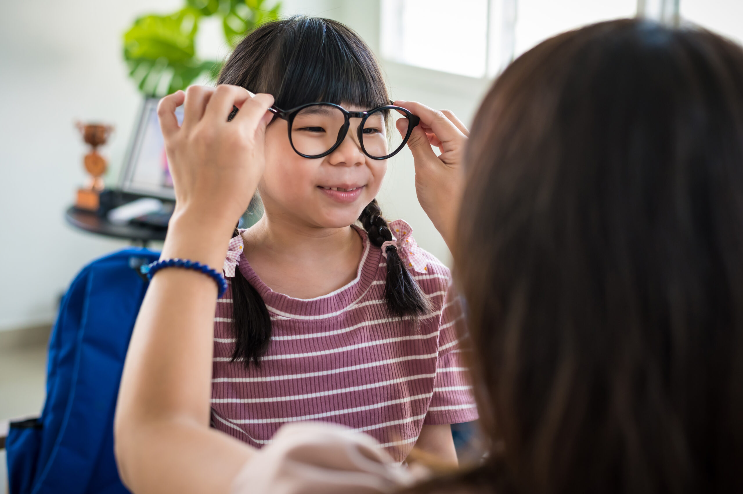 Young Asian mother putting eyeglasses on her daughter in living room and preparing her for school reopen day.